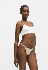 Γυναίκα με κοτσίδες, που φοράει λευκό αμάνικο crop top και μπεζ κάτω μέρος μπικίνι, στέκεται μπροστά σε ένα απλό ανοιχτό φόντο.