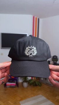 Fekete baseballsapka ívelt karimával, ezüst hímzett babérkoszorú díszítéssel és logóval az elején. Sima anyag finom textúrával.