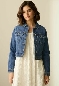 Chaqueta de mezclilla recortada en azul medio con herrajes plateados, dos bolsillos en el pecho y un cuello clásico. Llevada sobre un vestido blanco texturizado.