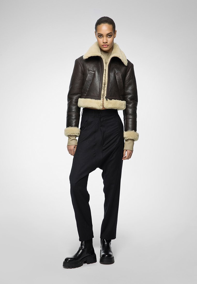 Dunkelbraune Lederjacke im Cropped-Stil mit cremefarbenem Kunstfellkragen und -manschetten, getragen über einem beigen Oberteil, kombiniert mit schwarzen hochtaillierten Hosen und Stiefeln.