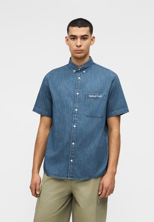 Jeune homme portant une chemise en denim bleu à manches courtes avec une poche poitrine et un pantalon cargo beige, debout devant un fond uni.