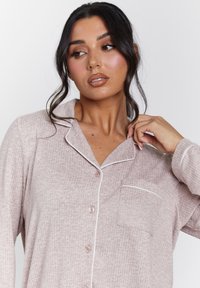 Camisa de pijama de color rosa claro, con canalé, cuello, cierre de botones y bolsillo frontal. Acentos de piping blanco alrededor del cuello y el bolsillo.
