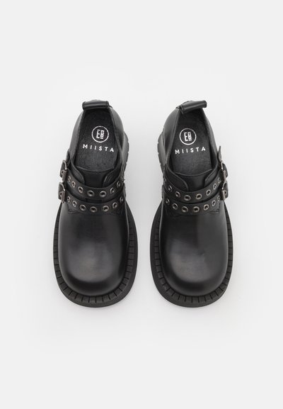 Pantofi negri din piele cu două curele ajustabile cu ochiuri din metal mari. Bot rotund și talpă din cauciuc texturată cu margine zimțată.