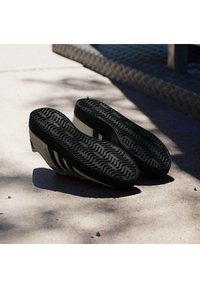 Crno-smeđe atletske cipele s teksturiranim gumenim potplatom, istaknutim utorima i tri crne pruge sa strane.