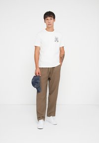 White short-sleeve T-shirt with a black graphic pocket, brown textured pants, and a dark blue cap held in hand, paired with white sneakers.