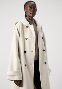 Lys beige dobbeltspent frakk med svarte knapper og brede mansjetter. Laget av myk materiale, med skrågcollar og rene linjer.