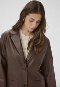 Junge Frau mit langen braunen Haaren schaut nach unten, trägt goldene Creolen, einen cremigen Strickpullover mit floralen Knöpfen und ein dunkelbraunes Lederblazer.
