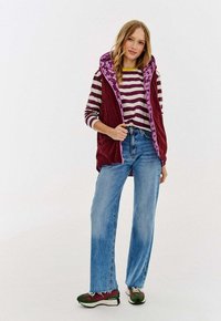 Kobieta w niebieskich dżinsach, w paskowanej koszulce z długim rękawem, bordowej kamizelce z fioletowymi kwiatowymi liniami i zielono-bordowych trampkach, ustawiona na tle bez wzorów.