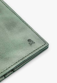 Esquina de billetera de cuero verde con textura detallada, costuras visibles y logo en relieve de calavera con huesos cruzados.