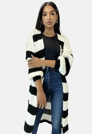 Kobieta o długich ciemnych włosach, nosząca złote kolczyki-koła, czarną bluzkę, niebieskie dżinsy oraz długi czarno-biały prążkowany sweter na drutach.