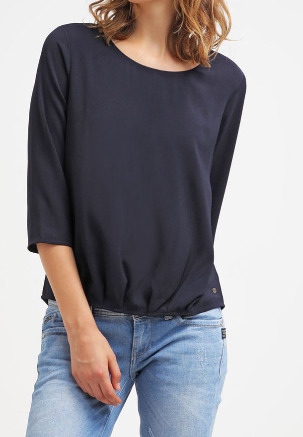 Femme portant un haut noir à manches trois-quarts et un jean bleu clair sur fond blanc uni.