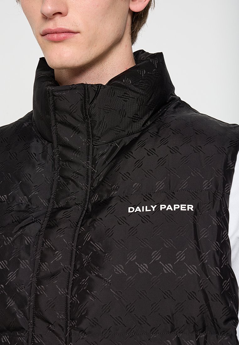 Man in een zwart gewatteerd vest met hoge kraag, subtiel reliëfpatroon en het "DAILY PAPER" logo op de borst.