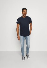 Tengerészkék színű rövid ujjú pamut póló, kerek nyakkivágással és fehér díszítéssel a karon. Világoskék, testhez simuló farmernadrággal és fekete sportcipővel párosítva.