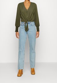 Blusa verde oliva con scollatura a V, chiusura frontale a bottoni e un fiocco in vita, abbinata a jeans dritti azzurri e stivali con tacco marroni.