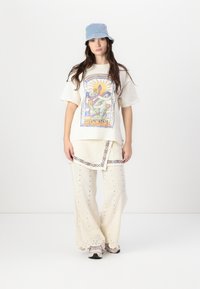 T-shirt oversize beige clair avec un design graphique coloré, associé à un pantalon large avec des détails en œillets et des accents brodés. Chapeau seau bleu.