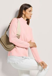 Frau mit großen Ohrhängern, pinkem Pullover, weißen Hosen und einer beige Umhängetasche, die seitlich vor einem hellen, einfarbigen Hintergrund posiert.