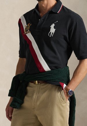 Homme portant un polo noir avec une bande diagonale rouge et blanche et un logo, un pantalon beige, un pull vert noué autour de la taille et une montre bleue au poignet.