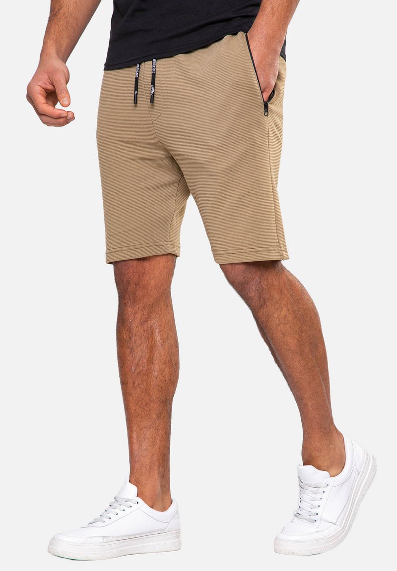 Shorts en mélange de coton beige avec un design nervuré texturé, dotés de poches zippées et de cordons de serrage contrastants, assortis à des baskets blanches.