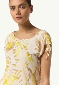 Bluse mit Blumenmuster aus weichem Stoff, ausgestattet mit kurzen Puffärmeln und Kordelzügen. Farben sind Pastellgelb und Beige.