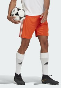 Orangene Fußballshorts mit drei weißen Streifen, kombiniert mit weißen Socken mit schwarzen Akzenten und schwarzen Fußballschuhen. Hält einen Fußball.