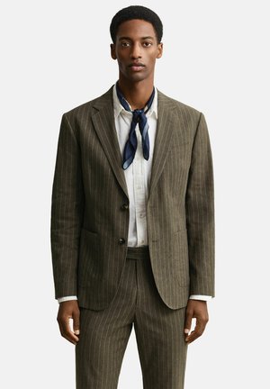 Jonge man die een bruin krijtstreep pak, wit overhemd en een marineblauwe sjaal om zijn nek draagt, staand tegen een effen achtergrond.