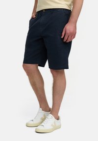 Shorts en coton navy avec poches latérales, fermeture éclair à l'avant et coupe décontractée. Associés à des baskets blanches avec des accents crème.