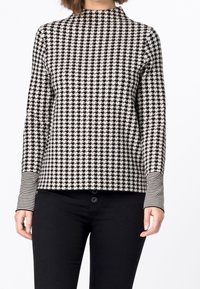 Woman wearing a black and white houndstooth patterned long-sleeve top with striped cuffs and black pants, against a white background.