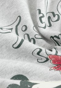 Tessuto di cotone grigio con una consistenza morbida, caratterizzato da testo corsivo verde e una grafica di fiore rosso, leggermente stropicciato e sovrapposto.