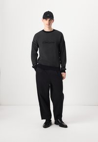Pulover negru din tricot cu textură, cu logo brodat, asortat cu pantaloni negri largi și pantofi negri, completat de o șapcă neagră.