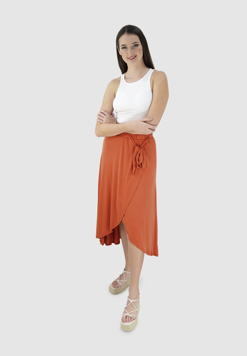 Wikkelrok in verbrande oranje stof, met een strikdetail in de taille en een vloeiende uitstraling. Gecombineerd met beige strap-sandalen van espadrilles.