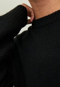 Pull noir en maille avec un col et des poignets côtelés. Le tissu semble doux et texturé, vue rapprochée sur le col et la manche.