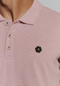 Polo shirt in lichtroze met een gestructureerde stof, drie knopen en een zwart hexagonaal logo met gouden accenten op de linker borst.