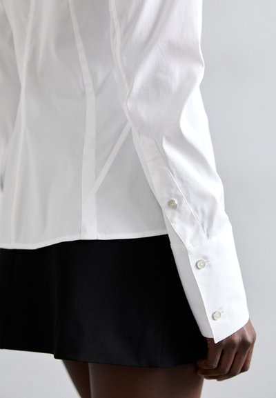 White cotton shirt with long sleeves, featuring buttoned cuffs and a fitted design, paired with a black skirt. Textured fabric visible.