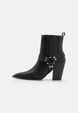 Sorte lær ankle boots med spiss tå, vertikale ribbede detaljer og et bredt belte med sølvfiner. Kraftig hæl med klar aksent.