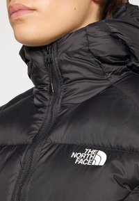 Zwarte pufferjas met een hoge kraag, ritssluiting en witte logo-detail op de linkerborst. Heeft een gladde, glanzende textuur.