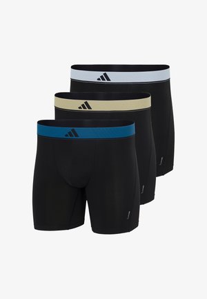 Mężczyzna bez koszulki, stojący boso, ubrany w czarne bokserki z szarym pasem w talii i logo Adidas na tle jednolitej białej ściany.