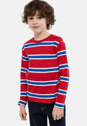 Garçon aux cheveux bouclés portant un t-shirt rouge à manches longues avec des rayures horizontales bleues et blanches et un jean foncé, debout devant un fond blanc.