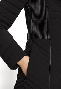 Czarna kurtka puchowa z przekątnym pikowaniem, błyszczącymi czarnymi akcentami w prążki, suwakiem z boku oraz paskiem w talii, co zapewnia dopasowany wygląd.