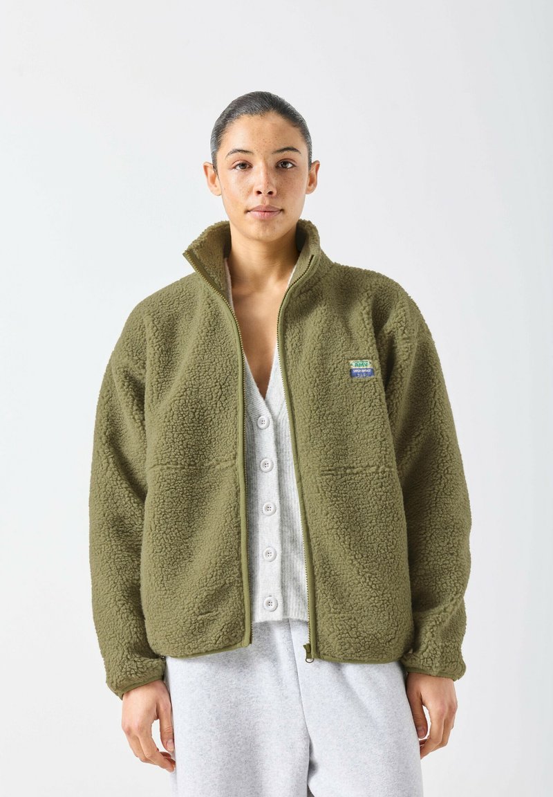 Ženska s temnimi lasmi nosi olično zeleno flis jakno čez bel zgornji del s srajčnimi gumbi in svetlo sive hlače na enostavni belem ozadju.