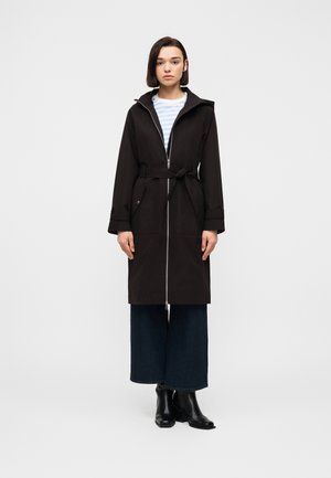 Jeune femme portant un manteau noir ceinturé avec une fermeture éclair à l'avant, un jean bleu à jambes larges, des bottines noires à talons et un haut rayé bleu clair.