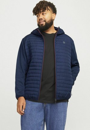 Marineblå isoleret jakke med quiltet front, fuld lynlåsdesign og hætte. Har elastiske manchetter og en solid sort inderside.