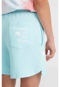 Helle blaue Shorts aus weichem Stoff, mit einer Gesäßtasche, die mit "Women of the Wave" bedruckt ist und einem weißen Akzent verziert ist. Der Saum ist leicht abgerundet.