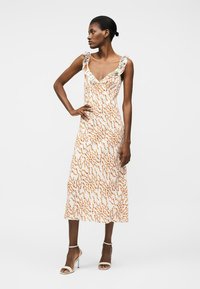 Mujer con un vestido midi sin mangas, blanco con patrones florales en naranja y verde y escote con volantes, combinado con sandalias blancas de tacón alto.