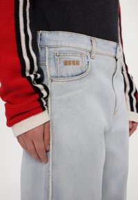 Personne portant un jean bleu clair avec le logo MSGM sur la poche avant et une manche de pull rouge, noire et blanche à rayures visible sur le bras gauche.