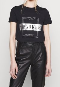 Camiseta de algodón negra con un gráfico metálico plateado de "SPARKLE" y texto debajo. Combinada con pantalones de cintura alta de cuero sintético negro.