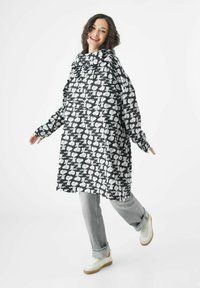 Schwarz-weißer gemusterter, übergroßer Mantel mit Kapuze, der ein verspieltes Design aufweist. Kombiniert mit hellgrauen Jeans und weißen Sneakern.