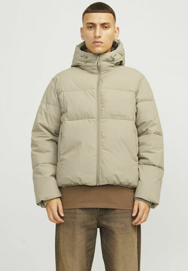Heledavah beige värviga paks jakk kapuutsiga, millel on esine lukuga, elastsed mansetid ja lühike lõige. Kandukas pruuni pika varrukaga särgi üle.