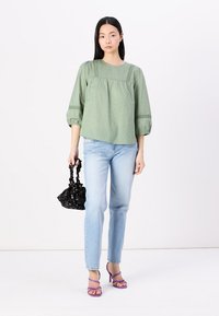 Blusa verde con texture e maniche a sbuffo, abbinata a jeans azzurri. Una borsa nera e sandali con tacco viola completano l'outfit.
