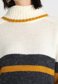 Strickpullover mit hohem Kragen, cremefarbener Basis, dunkelgrauen und senffarbenen Streifen sowie einer strukturierten Oberfläche mit sichtbaren Nahtdetails.