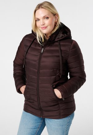 Vrouw met blond haar, die grote oorbellen draagt en een donkerbruin gewatteerd jack met capuchon heeft, met haar handen in haar zakken, staand tegen een lichte achtergrond.